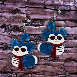 May include: A pair of whimsical earrings featuring a cartoon worm design. Each earring has a white body with black stripes, a red scarf, and blue spiky hair. The worm wears sunglasses and has a glittery finish.