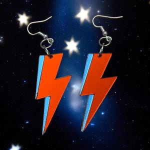 May include: A pair of earrings featuring a red lightning bolt design with a light blue outline. The earrings are suspended from silver-colored hooks. The background is a dark blue space scene with stars.