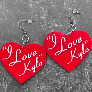 Puede incluir: Un par de pendientes de corazón rojo con ganchos plateados. Los pendientes tienen el texto "I Love Kylo" escrito en blanco.