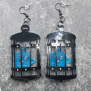 Puede incluir: Un par de pendientes de metal negro con un fantasma azul atrapado dentro de una jaula. Los pendientes cuelgan de ganchos negros.