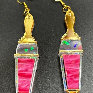 May include: A pair of gold-tone earrings with a pink and iridescent glass design. The earrings are shaped like a bottle of lipstick.