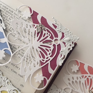 May include: A white paper butterfly bookmark with a delicate cut-out design. The bookmark is placed on a book with a pink cover and white text. The butterfly has a small, clear, sparkly embellishment on its wing.
