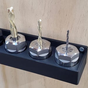 Puede incluir: Un soporte negro para brocas con cuatro brocas. El soporte es rectangular y está montado sobre una superficie de madera de color claro. Las brocas son de color plateado y dorado.