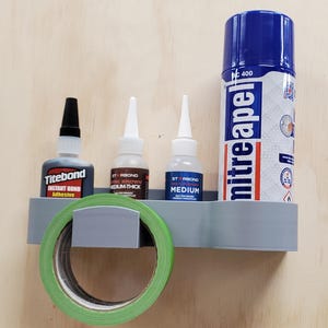 May include: A gray wall-mounted organizer with three bottles of glue and a roll of green tape. The bottles are labeled "Titebond Instant Bond Adhesive", "Starbond Dark Brown Medium-Thick", and "Starbond Medium". The spray can is labeled "Mitreapel 400".