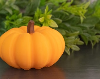 May include: A small, orange pumpkin with a brown stem. The pumpkin has vertical ridges and sits on a dark surface, with green foliage in the background. A seasonal decoration.