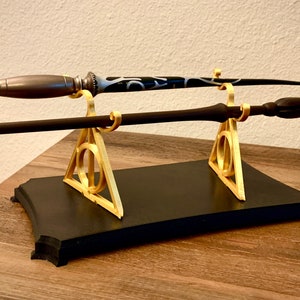 May include: Two wands are displayed on a black stand with gold-colored supports. The wands have unique designs, one with a dark handle and the other with a silver and black handle. The stand features a Deathly Hallows symbol.
