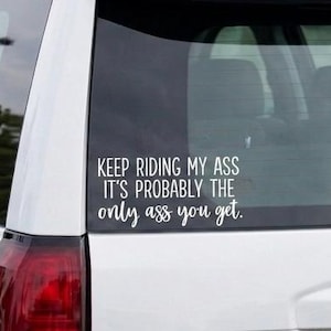 May include: A white SUV features a white vinyl decal on the rear window with the text "KEEP RIDING MY ASS IT'S PROBABLY THE only ass you get." The decal uses a combination of bold block lettering and a cursive script font for the final word.