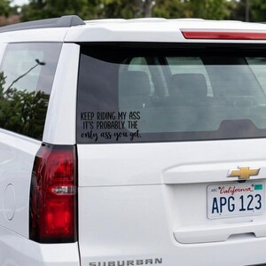 May include: A white SUV with a black vinyl decal applied to the rear window. The decal features black text that reads: KEEP RIDING MY ASS IT'S PROBABLY THE only ass you get. The vehicle has a California license plate and red taillights.