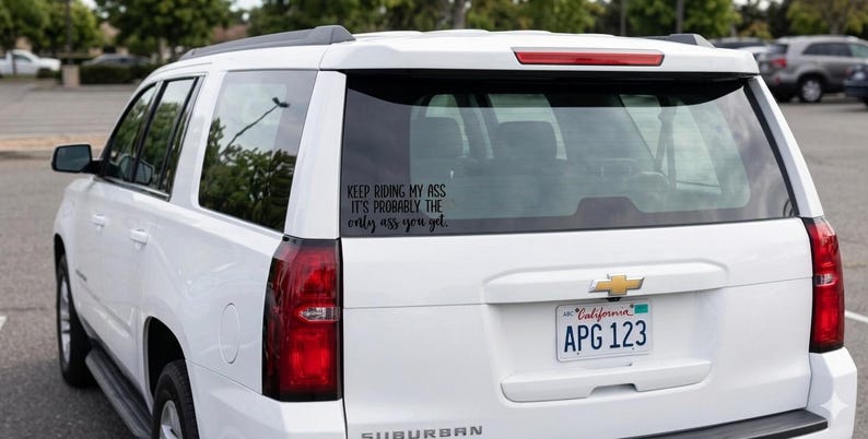 May include: A white SUV with a black vinyl decal applied to the rear window. The decal features black text that reads: KEEP RIDING MY ASS IT'S PROBABLY THE only ass you get. The vehicle has a California license plate and red taillights.