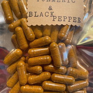 May include: A clear plastic bag filled with numerous turmeric and black pepper capsules. The capsules are a golden-brown color and are labeled with a brown paper tag that reads "TURMERIC & BLACK PEPPER".