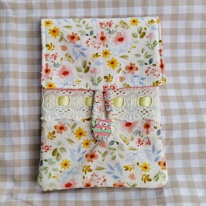 May include: A floral print case with a flap closure. The case has a white background with pink, yellow, and orange flowers, green leaves, and blue bows. A decorative lace trim with yellow ribbon and an owl button are also visible.