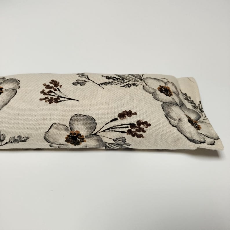 Op de afbeelding: Een wit oogkussen met een bloemenpatroon met grijze en bruine bloemen. Het kussen is gemaakt van een zachte, natuurlijke stof.
