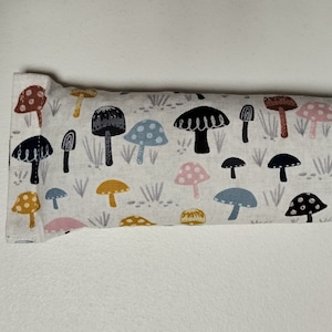 Op de afbeelding: Een wit stoffen oogkussen met een kleurrijke paddenstoelprint. De paddenstoelen zijn in verschillende kleuren, waaronder roze, blauw, geel en bruin. Het kussen is rechthoekig en heeft een naad in het midden.