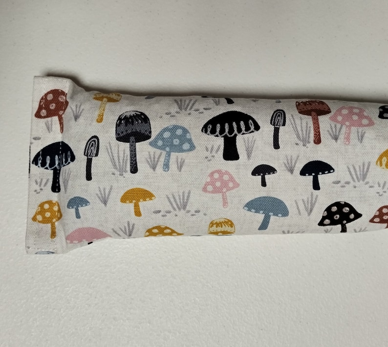 Op de afbeelding: Een wit stoffen oogkussen met een kleurrijke paddenstoelprint. De paddenstoelen zijn in verschillende kleuren, waaronder roze, blauw, geel en bruin. Het kussen is rechthoekig en heeft een naad in het midden.