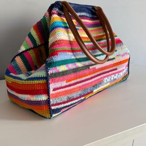 Op de afbeelding: Een grote, kleurrijke gehaakte tote bag met een verscheidenheid aan horizontale strepen in tinten rood, roze, oranje, groen, blauw en geel. De tas heeft bruine leren handvatten en een donkerblauwe bovenkant.