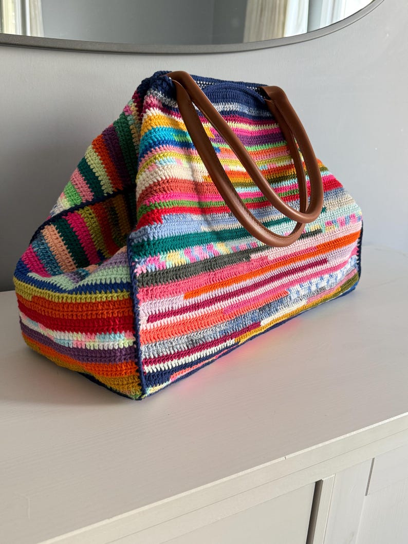 Op de afbeelding: Een grote, kleurrijke gehaakte tote bag met een verscheidenheid aan horizontale strepen in tinten rood, roze, oranje, groen, blauw en geel. De tas heeft bruine leren handvatten en een donkerblauwe bovenkant.