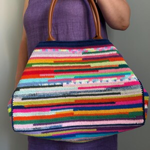 Op de afbeelding: Een grote, kleurrijke gehaakte tote bag met een gestreept patroon in verschillende kleuren, waaronder rood, geel, blauw en roze. De tas heeft bruine leren handvatten en een donkerblauwe bovenrand. De tas wordt door een persoon vastgehouden.