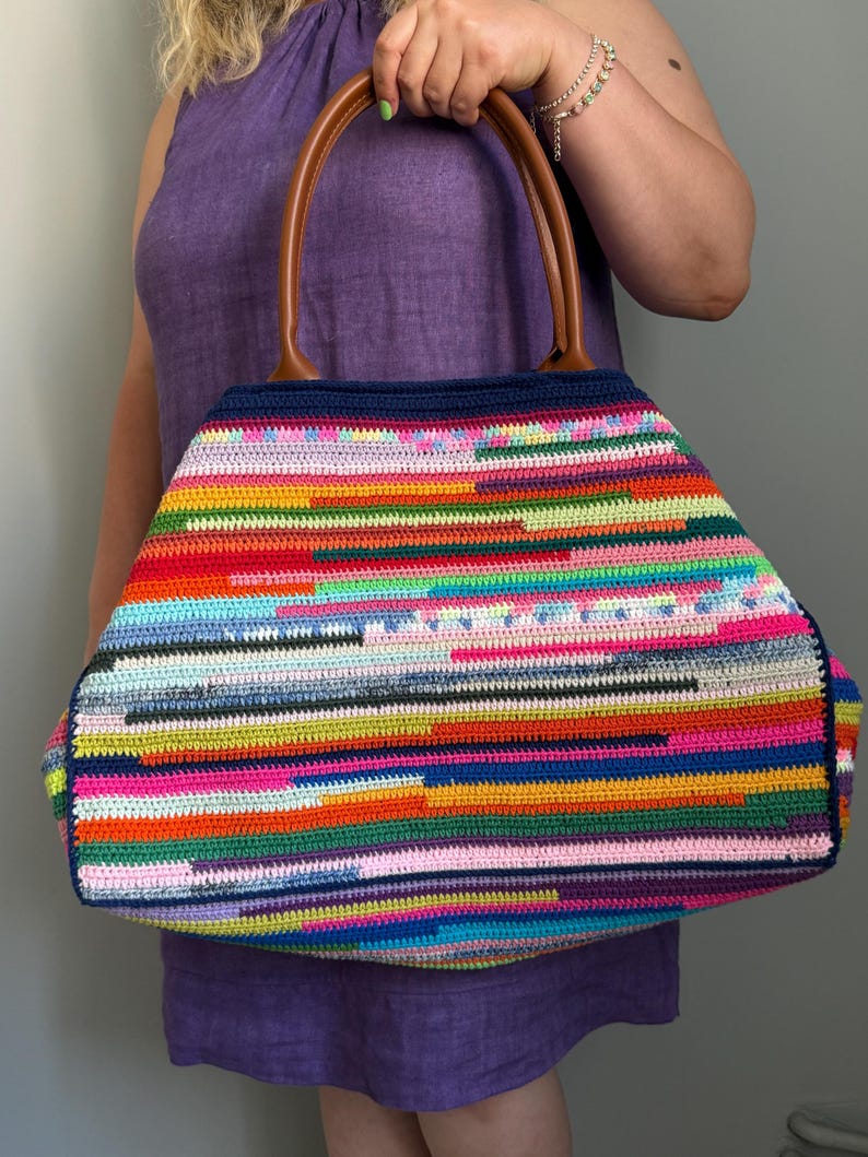 Op de afbeelding: Een grote, kleurrijke gehaakte tote bag met een gestreept patroon in verschillende kleuren, waaronder rood, geel, blauw en roze. De tas heeft bruine leren handvatten en een donkerblauwe bovenrand. De tas wordt door een persoon vastgehouden.