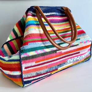 Op de afbeelding: Een grote, kleurrijke gehaakte tote bag met bruine leren handvatten. De tas heeft horizontale strepen in verschillende kleuren, waaronder rood, oranje, geel, groen, blauw en roze. De tas is aan de bovenkant open.