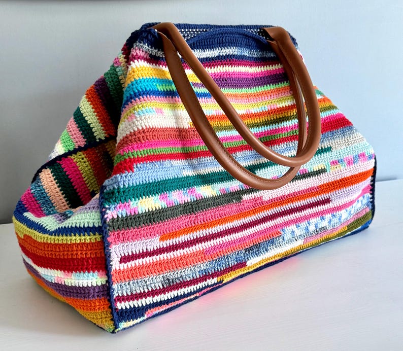 Op de afbeelding: Een grote, kleurrijke gehaakte tote bag met bruine leren handvatten. De tas heeft horizontale strepen in verschillende kleuren, waaronder rood, oranje, geel, groen, blauw en roze. De tas is aan de bovenkant open.