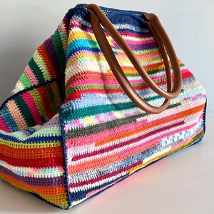 Op de afbeelding: Een grote, handgemaakte gehaakte tote bag met een levendig, veelkleurig gestreept patroon. De tas heeft een marineblauwe bovenkant en bruine leren handvatten. De tas is gemaakt van verschillende kleuren, waaronder roze, groen, oranje en paars.