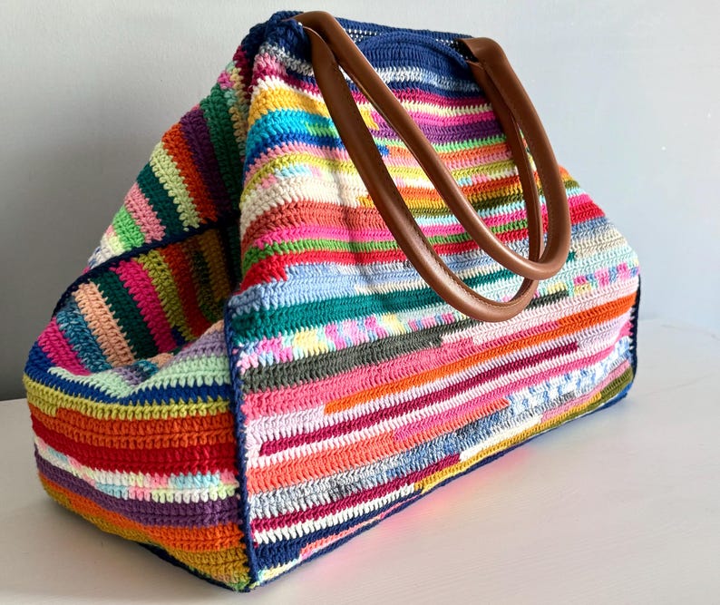 Op de afbeelding: Een grote, handgemaakte gehaakte tote bag met een levendig, veelkleurig gestreept patroon. De tas heeft een marineblauwe bovenkant en bruine leren handvatten. De tas is gemaakt van verschillende kleuren, waaronder roze, groen, oranje en paars.