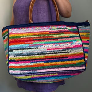 Op de afbeelding: Een grote, kleurrijke gehaakte tote bag met bruine leren handvatten. De tas heeft een levendig, horizontaal gestreept patroon in verschillende kleuren, waaronder rood, blauw, groen en roze. De bovenrand is donkerblauw. De tas wordt gedragen door een persoon die een paarse jurk draagt.