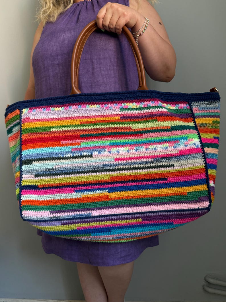 Op de afbeelding: Een grote, kleurrijke gehaakte tote bag met bruine leren handvatten. De tas heeft een levendig, horizontaal gestreept patroon in verschillende kleuren, waaronder rood, blauw, groen en roze. De bovenrand is donkerblauw. De tas wordt gedragen door een persoon die een paarse jurk draagt.