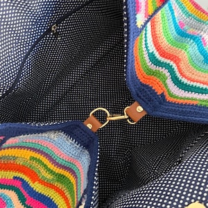 Op de afbeelding: Een kleurrijke, handgemaakte gehaakte tote bag. De tas heeft een levendig regenboogpatroon aan de buitenkant, met een marineblauwe rand en accenten. De binnenkant is gevoerd met marineblauwe stof met witte stippen. Het heeft een gouden sluiting en bruine leren bandjes.