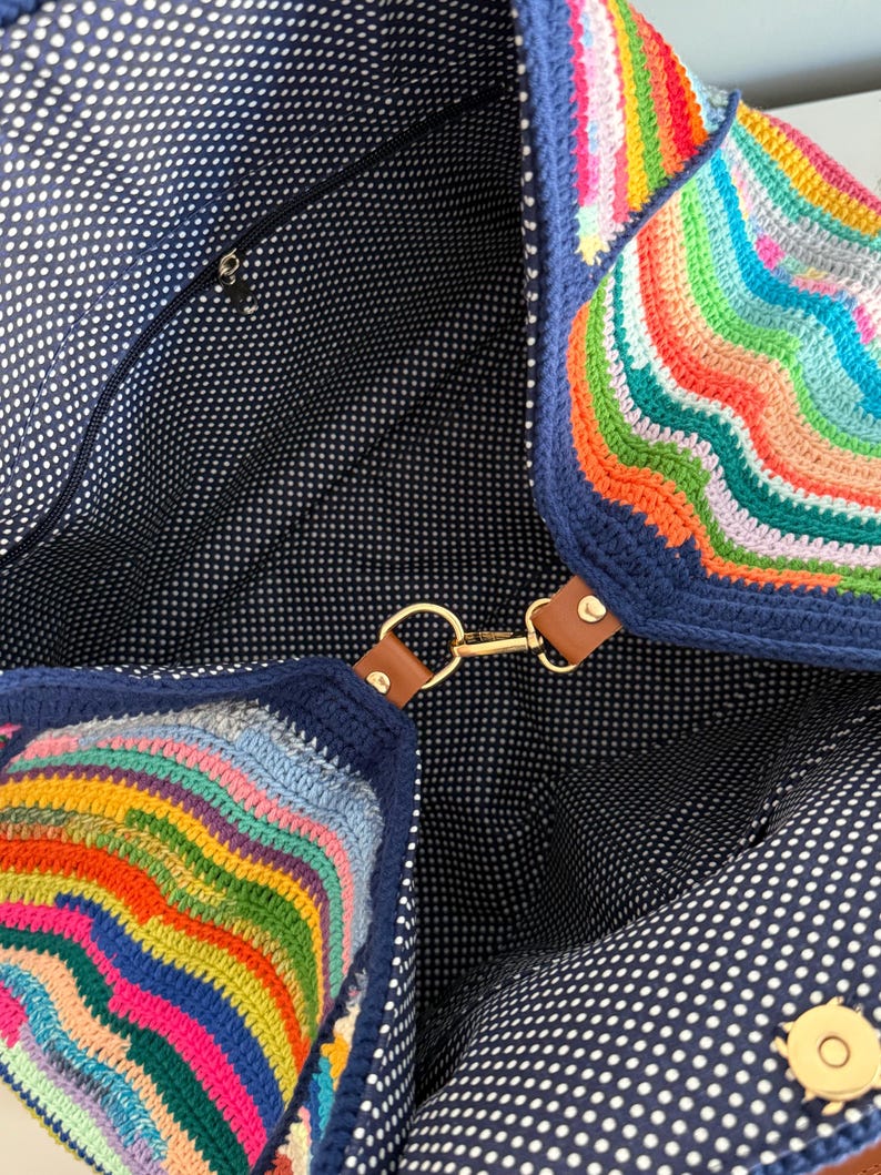 Op de afbeelding: Een kleurrijke, handgemaakte gehaakte tote bag. De tas heeft een levendig regenboogpatroon aan de buitenkant, met een marineblauwe rand en accenten. De binnenkant is gevoerd met marineblauwe stof met witte stippen. Het heeft een gouden sluiting en bruine leren bandjes.