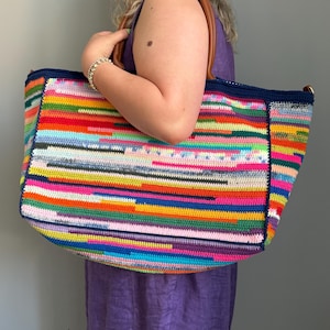 Op de afbeelding: Een grote, rechthoekige gehaakte tote bag met een levendig, veelkleurig gestreept patroon. De tas heeft bruine leren handvatten en een marineblauwe rand. De tas wordt vastgehouden door een persoon die een paarse jurk draagt.