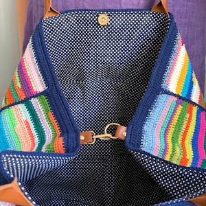 Op de afbeelding: Een kleurrijke gehaakte tote bag met een marineblauwe rand en bandjes. De tas heeft een levendig patchwork-ontwerp met verschillende kleuren. De binnenkant is gevoerd met een marineblauwe stof met witte stippen. De tas heeft een magnetische druksluiting en een goudkleurige sluiting.