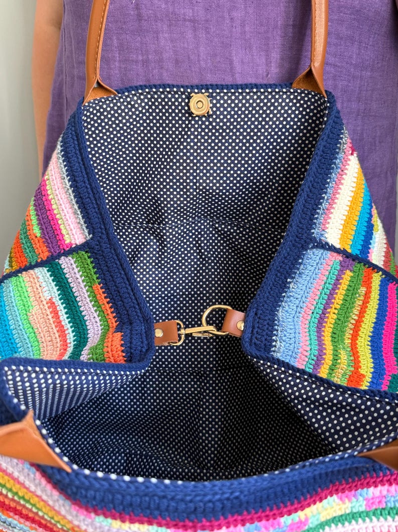 Op de afbeelding: Een kleurrijke gehaakte tote bag met een marineblauwe rand en bandjes. De tas heeft een levendig patchwork-ontwerp met verschillende kleuren. De binnenkant is gevoerd met een marineblauwe stof met witte stippen. De tas heeft een magnetische druksluiting en een goudkleurige sluiting.
