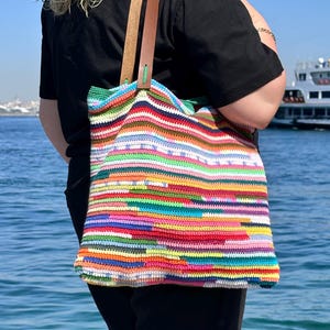 May include: A colorful, handmade crochet tote bag with brown leather handles. The bag features a vibrant mix of horizontal stripes in various colors, including red, green, blue, and yellow. The bag is held by a person wearing a black shirt.