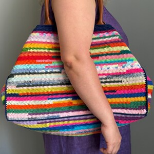 Op de afbeelding: Een grote, kleurrijke gehaakte tote bag met een regenboog gestreept patroon. De tas heeft bruine leren handvatten en een marineblauwe rand. De tas wordt vastgehouden door een persoon die een paarse jurk draagt.
