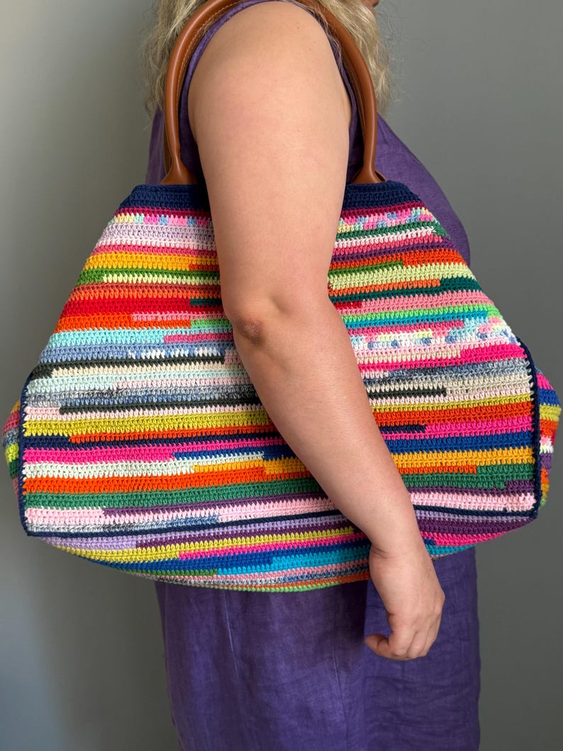 Op de afbeelding: Een grote, kleurrijke gehaakte tote bag met een regenboog gestreept patroon. De tas heeft bruine leren handvatten en een marineblauwe rand. De tas wordt vastgehouden door een persoon die een paarse jurk draagt.