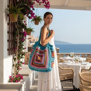 May include: A turquoise crochet tote bag with a colourful square pattern. The bag features a large central square with pink, orange, and yellow details, surrounded by a blue border. The bag is carried over the shoulder.