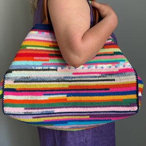 Op de afbeelding: Een grote, kleurrijke gehaakte draagtas met een verscheidenheid aan horizontale strepen in tinten rood, oranje, geel, groen, blauw, roze en paars. De tas heeft bruine leren handvatten en een donkerblauwe rand.
