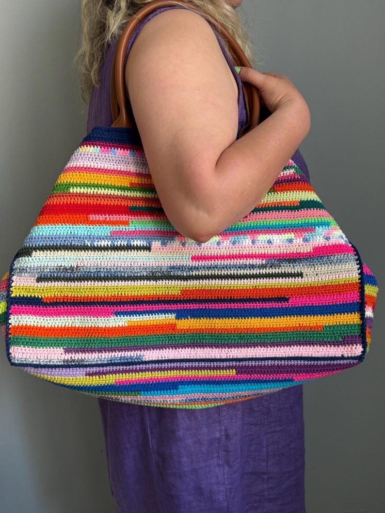 Op de afbeelding: Een grote, kleurrijke gehaakte draagtas met een verscheidenheid aan horizontale strepen in tinten rood, oranje, geel, groen, blauw, roze en paars. De tas heeft bruine leren handvatten en een donkerblauwe rand.