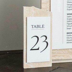 May include: A bar menu and table number 23 displayed on a wooden table. The bar menu lists signature drinks, liquor, wine, and beer. The table number is in a black font on a white background.