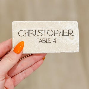 May include: A rectangular, off-white place card with the name "CHRISTOPHER" and "TABLE 4" printed in black. The card has a textured surface and is held by a hand with orange nail polish.