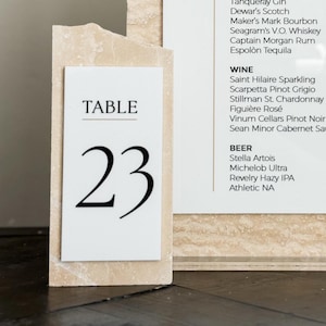 May include: A bar menu and table number 23 are displayed on a light beige stone-like background. The bar menu lists signature drinks, liquor, wine, and beer. The table number is a white card with the number 23 in a large, bold font.