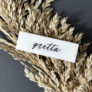 May include: A white card with the name "gretta" written in black cursive script. The card is lying on top of a bunch of dried beige grass.