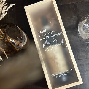 May include: A wooden box with a frosted glass window, containing a bottle of wine. The box has a white inscription that reads "Pairs well with a new home. Place for us to drink. Congrats on the new place!"