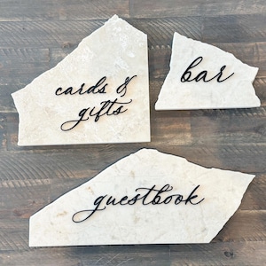 May include: Three irregular, cream-coloured stone slabs with rough, jagged edges. Each slab features black, cursive lettering. The signs read "cards & gifts," "bar," and "guestbook" in a flowing, elegant script style applied to the stone surface.