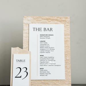 May include: A bar menu and table number 23 on a light-colored stone background. The bar menu lists signature drinks, liquor, wine, and beer. The table number is in a simple, modern font.