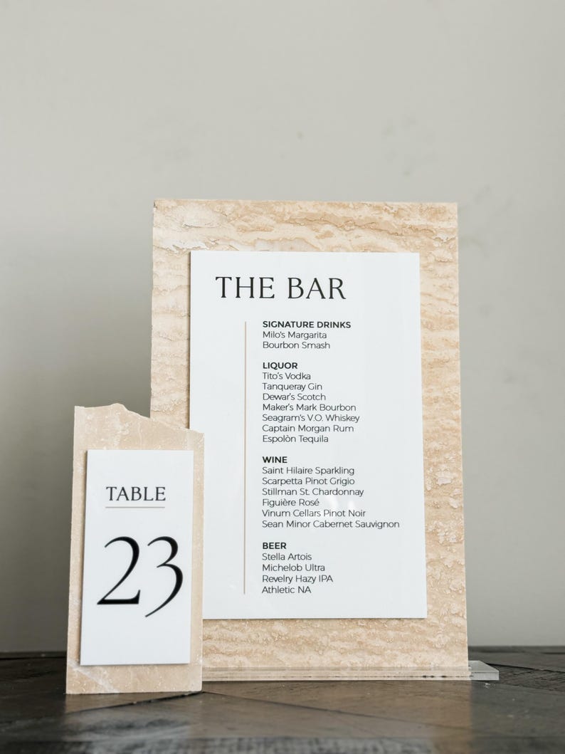 May include: A bar menu and table number 23 on a light-colored stone background. The bar menu lists signature drinks, liquor, wine, and beer. The table number is in a simple, modern font.