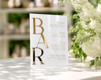 May include: A white rectangular acrylic bar menu sign featuring large, vertical, gold-toned reflective letters spelling BAR on the left side. The right side lists drink categories including Liquor, Wine, Beer, and Non-Alcoholic with specific beverage options printed in a gold-toned font.