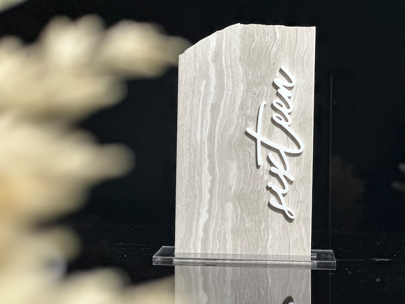 Op de afbeelding: Een tafelnummer met grijze marmertextuur en het woord "zestien" in een wit scriptlettertype. Het tafelnummer wordt weergegeven op een heldere acryl basis, geschikt voor evenementen en feesten.