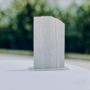 Op de afbeelding: Een rechthoekig, lichtgrijs houten tafelnummer met een gebogen bovenrand, geplaatst op een heldere acrylbasis. Het tafelnummer heeft een textuur van houtnerf. De achtergrond is wazig, met groene bladeren.